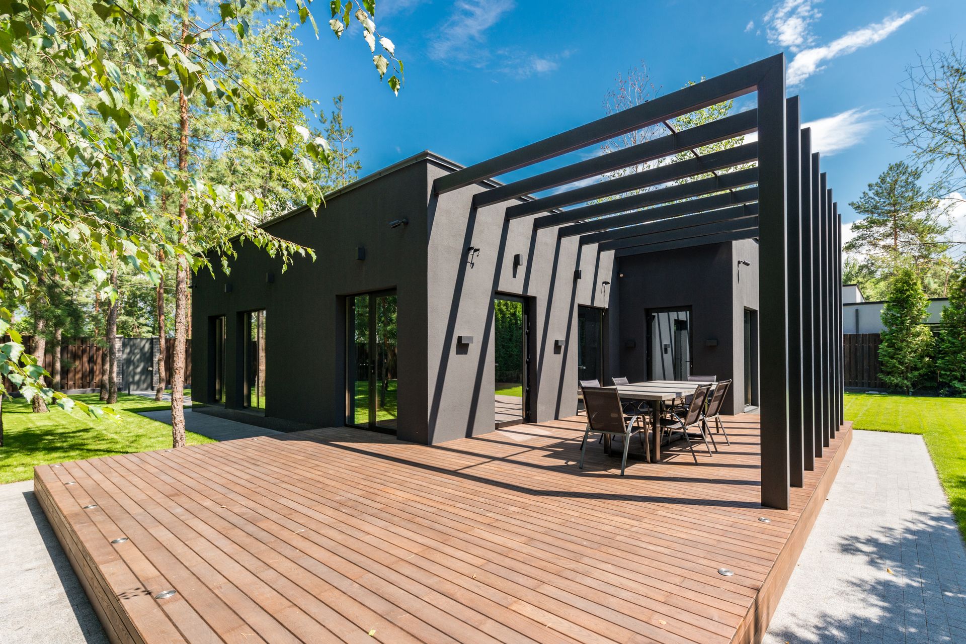 A custom wood deck with a black steel pergola and patio dining behind a modern home, framed by mature trees
